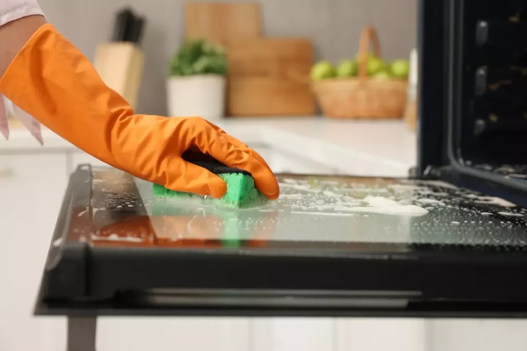 Woman cleaning oven door with sponge in kitchen.